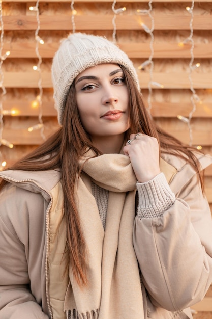 Retrato de una chica joven y elegante con una ropa exterior beige de moda con un sombrero de punto, un suéter clásico, una chaqueta y una bufanda cerca de una pared de madera con luces al aire libre