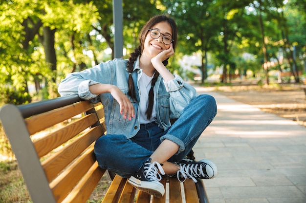 Retrato de una chica joven estudiante linda feliz complacida con anteojos sentado en un banco al aire libre en el parque natural.