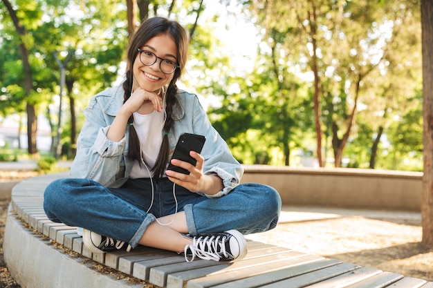 Retrato de una chica joven estudiante linda feliz positiva complacida con anteojos sentado en un banco al aire libre en el parque natural con teléfono móvil charlando escuchando música con auriculares.