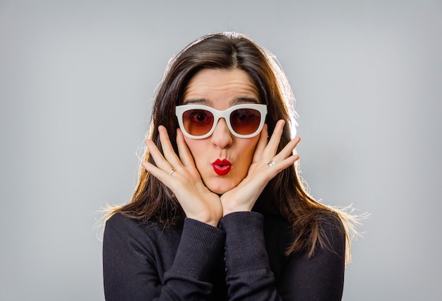 Retrato de chica joven real con gafas de sol blancas gesticulando con sus labios rojos sobre un fondo gris