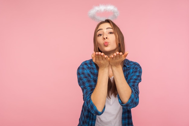 Retrato de una chica linda y encantadora con un halo angelical sobre la cabeza que sopla un beso de aire sensual a la cámara, coquetea y expresa amor, sentimientos románticos amorosos. tiro de estudio interior aislado sobre fondo de color rosa