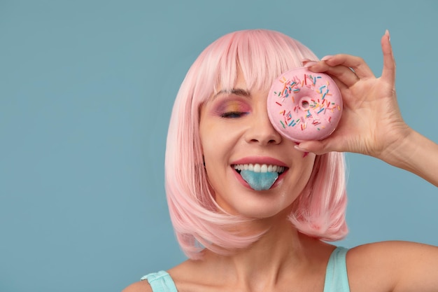 Retrato de una chica de moda con una peluca rosa y una rosquilla rosa en las manos que se mantiene a la vista muestra la lengua