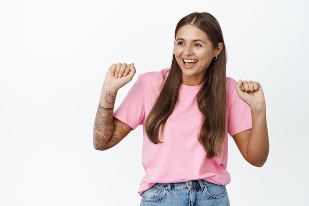 Retrato de chica moderna bailando, tiene tatuajes en la mano y piercing, sonriendo y riendo alegre, de pie en blanco