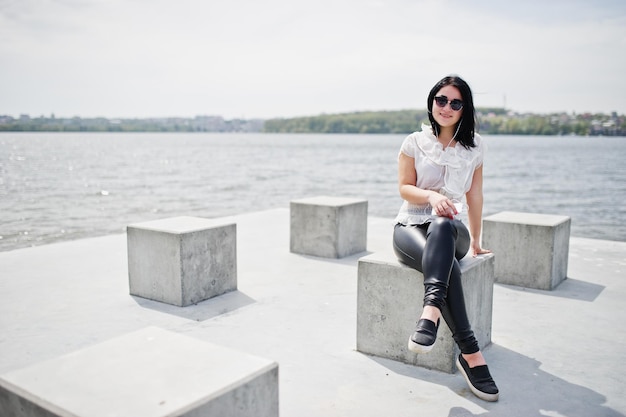 Retrato de una chica morena escuchando música en los auriculares del teléfono móvil en pantalones de cuero de mujer y gafas de sol de blusa blanca contra cubos de piedra en la playa del lago