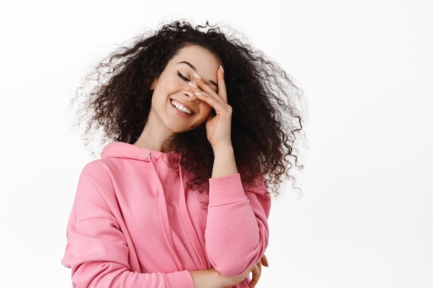 Retrato de una chica morena natural que cierra los ojos soñadora, toca la piel limpia y brillante y sonríe feliz, parada despreocupada contra el fondo blanco con capucha rosa