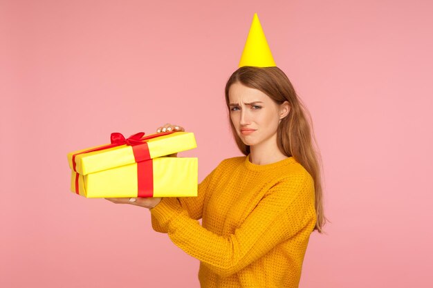 Retrato de una chica pelirroja infeliz con un cono parcial en la cabeza abriendo una caja de regalo y mirando tristemente a la cámara, decepcionada con la sorpresa de cumpleaños, mal regalo y vacaciones estropeadas. foto de estudio fondo rosa