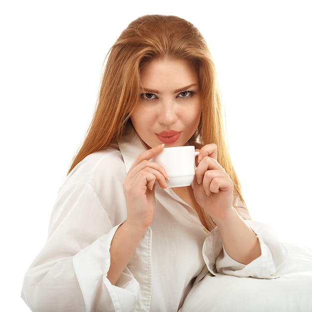 Retrato de una chica pelirroja sexy tomando café en la cama aislada en blanco