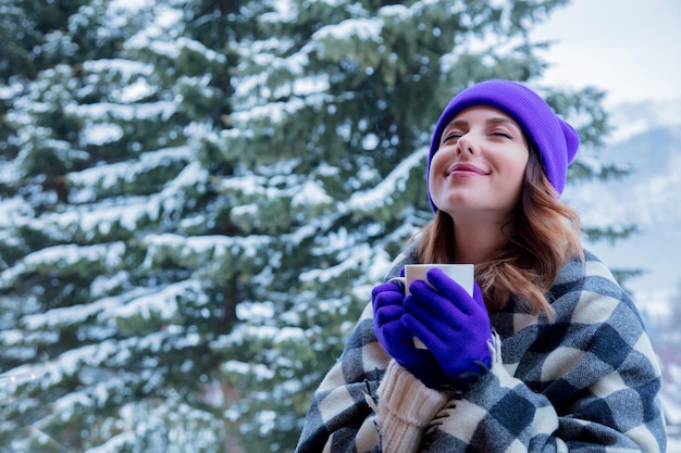 retrato de una chica pelirroja con sombrero a cuadros y púrpura con una taza de bebida caliente en el exterior de invierno