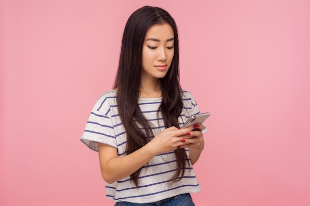 Retrato de una chica con el pelo moreno en una camiseta escribiendo un mensaje en un teléfono móvil leyendo una publicación en una red social chateando en línea con una expresión seria en un estudio interior aislado en un fondo rosa