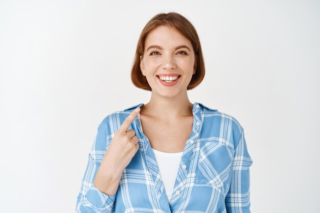 Retrato de una chica profesional sonriente apuntándose a sí misma autopromocionándose hablando de logros personales de pie con ropa informal de fondo blanco