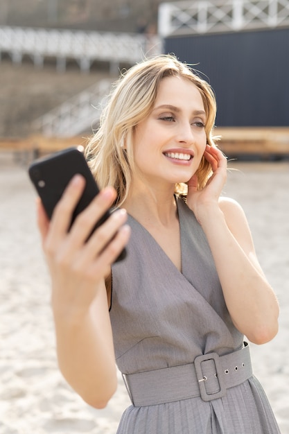 Foto retrato de una chica rubia alegre escuchando música y sosteniendo su teléfono móvil con una sonrisa en una playa