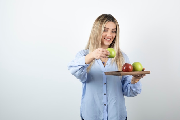 Retrato de una chica rubia sosteniendo una tabla de madera con manzanas verdes y rojas