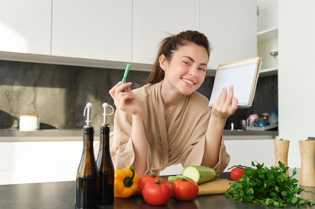 Retrato de una chica sonriente y guapa con una lista de compras sosteniendo un cuaderno y leyendo recetas de cocina