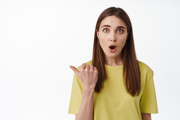 Retrato de una chica sorprendida apuntando a la izquierda, mirando con curiosidad a la cámara, preguntando sobre la promoción, intrigada con la publicidad, de pie contra el fondo blanco