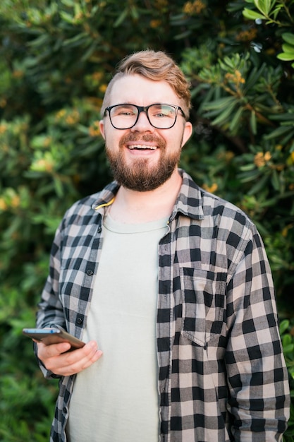 Retrato de un chico alegre y atractivo que usa un teléfono inteligente para desplazarse en las redes sociales sobre el fondo del árbol verde