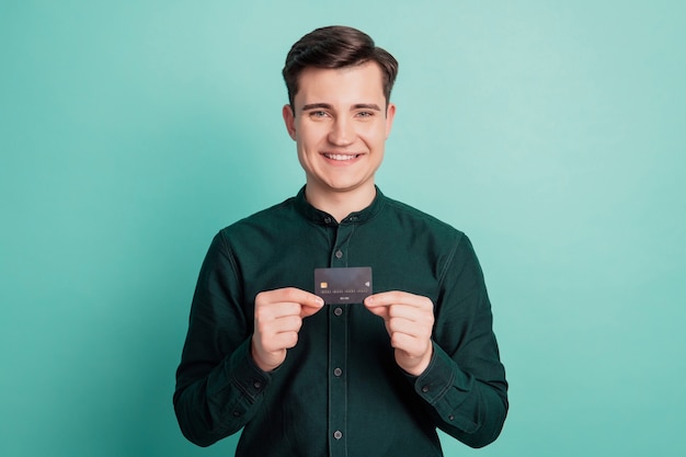 Retrato de chico alegre positivo mantenga tarjeta de crédito plástica posando sobre fondo turquesa