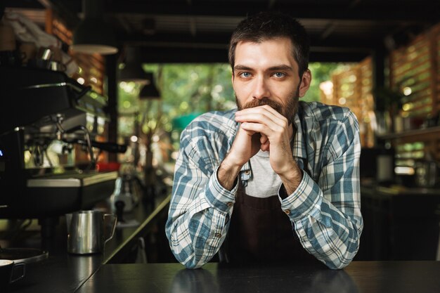 Retrato de chico guapo barista con delantal sonriendo mientras trabaja en la cafetería de la calle o al aire libre