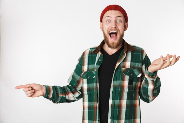 Retrato de chico hipster con sombrero y camisa a cuadros riendo y señalando con el dedo a un lado, mientras está de pie aislado sobre fondo blanco.