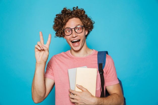 Retrato de chico joven inconformista con cabello rizado con gafas y mochila sonriendo y sosteniendo libros