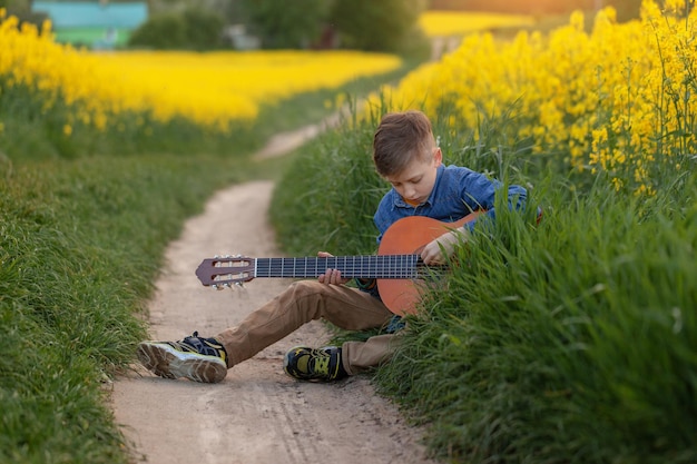 Retrato de un chico lindo tocando una guitarra sentado en la carretera en el campo amarillo de verano.
