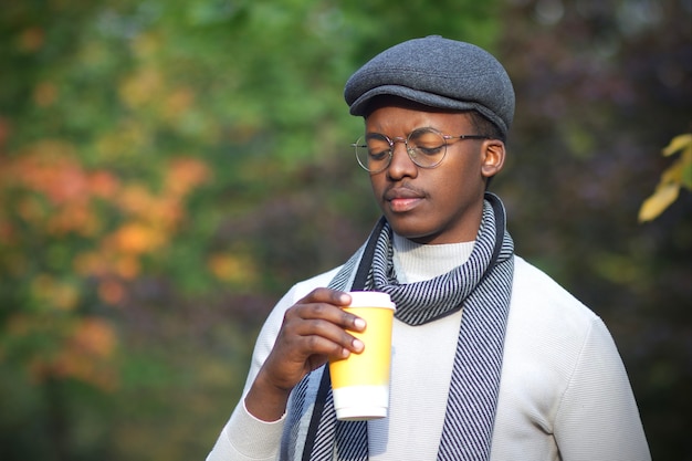 Retrato de chico negro triste deprimido frustrado molesto, apuesto joven afroamericano caminando en el parque de otoño dorado con sombrero, bufanda y vasos con taza de café, sintiéndose mal, infeliz