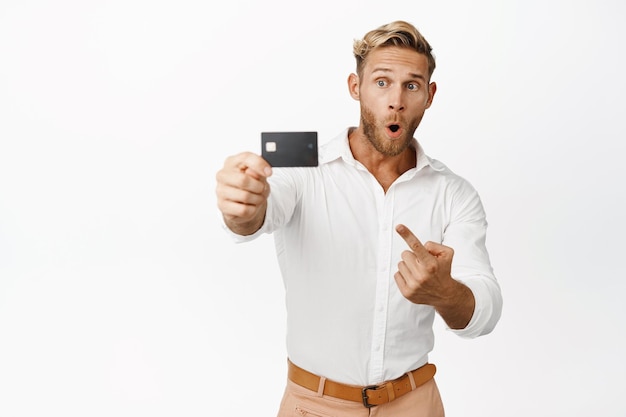 Retrato de chico rubio señalando y mostrando tarjeta de crédito demostrando oferta promocional de compras fondo blanco Copiar espacio