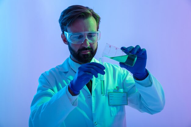 Retrato de un científico hombre confiado con unifrom y gafas que se encuentran aisladas sobre neblina azul-rosa, experimentando con tubos