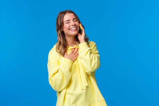 Retrato en la cintura de una alegre joven sonriente que tiene una conversación divertida en el teléfono, sosteniendo el teléfono inteligente cerca de la oreja, cierra los ojos y se ríe mientras toca el cofre, de pie sobre una pared azul