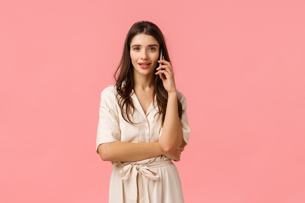 Retrato de cintura alta seria y hermosa mujer joven descarada vestida, sosteniendo el teléfono, hablando por teléfono, llamando a un amigo, conversando, verificando el pedido, de pie en la pared rosa