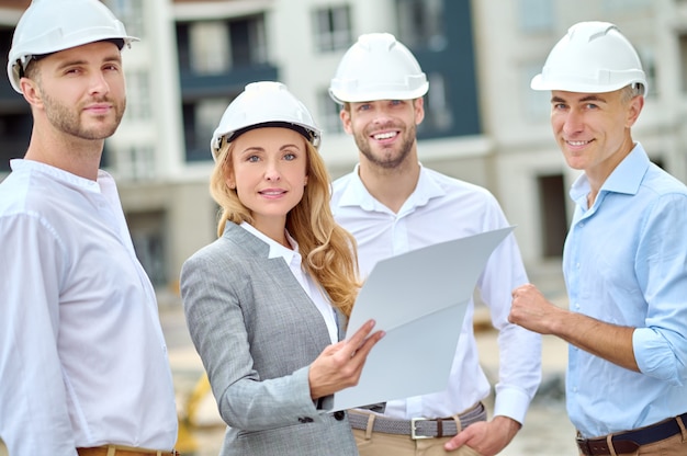 Retrato de cintura para arriba de una hermosa rubia tranquila confiada supervisora y trabajadores de la construcción en cascos de pie al aire libre