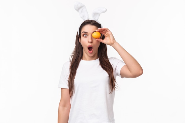 Retrato de cintura para arriba de una mujer joven asombrada y sorprendida que ve algo increíble, sostenga el huevo de color sobre el ojo, mirando a la cámara sorprendida, jadeando impresionado