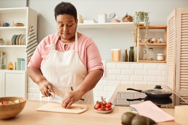 Retrato de cintura arriba en tonos cálidos de una anciana negra cortando verduras mientras hace ensalada en un kit acogedor