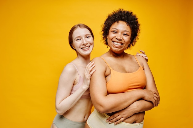 Retrato en la cintura de dos mujeres jóvenes sonrientes de piel oscura y piel clara abrazándose mientras están de pie de nuevo