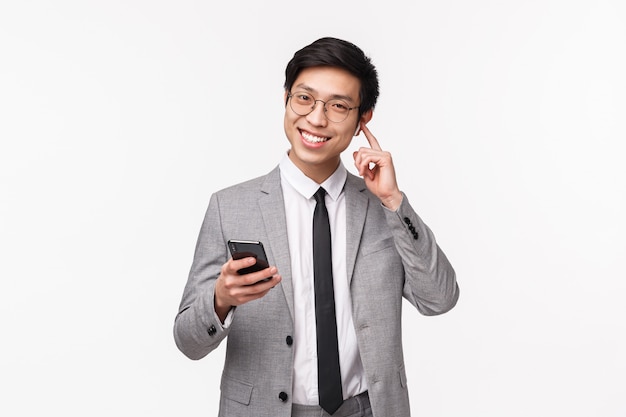 Retrato en la cintura del hombre de negocios asiático feliz satisfecho y sonriente con auriculares inalámbricos, sosteniendo el teléfono móvil, cambiar el volumen o llamar a alguien con auriculares, de pie sobre una pared blanca