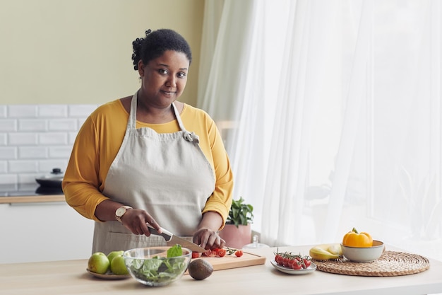 Retrato en la cintura de una joven negra cocinando una ensalada saludable en la cocina y mirando la copia de la cámara