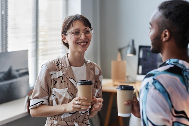 Retrato en la cintura de una joven sonriente disfrutando de un café para llevar mientras conversa con un compañero de trabajo en el trabajo