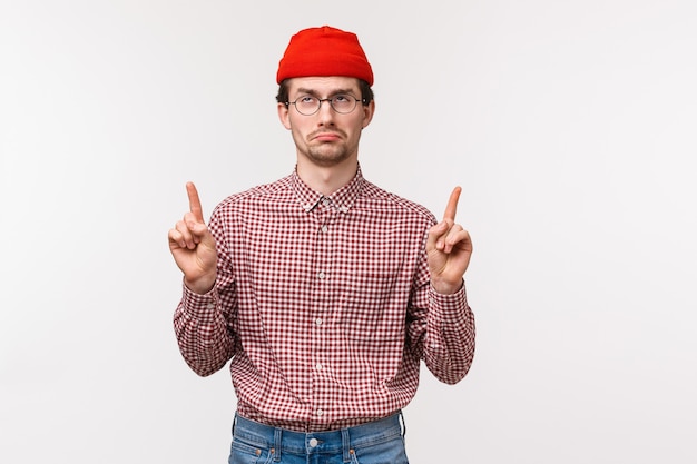 Foto retrato en la cintura molesto y decepcionado quejumbroso hombre adulto con gorro rojo, gafas, apuntando con los dedos hacia el anuncio y mostrando disgusto y sentimientos incómodos con expresión de la cara, pared blanca