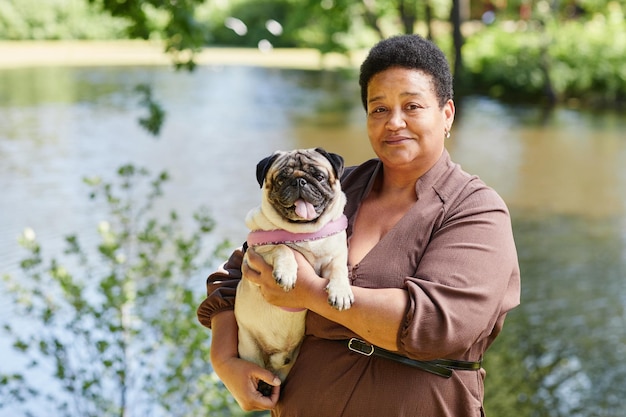 Retrato en la cintura de una mujer negra madura sosteniendo un lindo perro pug y mirando a la cámara en un hermoso parque