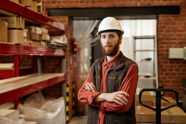 Retrato en la cintura de un trabajador barbudo que usa casco en el almacén de la fábrica y mira la cámara mientras