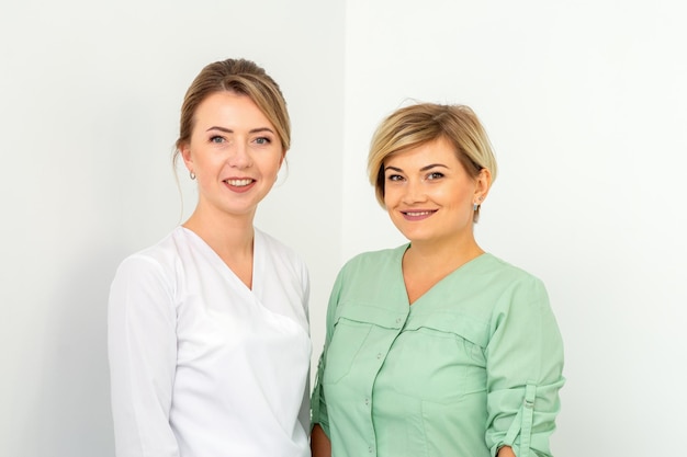 Retrato de clausura de dos jóvenes trabajadoras sanitarias caucásicas sonrientes de pie mirando a la cámara sobre fondo blanco.