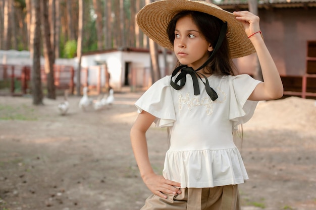 Retrato de clausura de morena preadolescente enfocada en blusa ligera y sombrero de paja de ala grande de pie en el patio de la casa de campo ubicada en un bosque de pinos mirando hacia otro lado