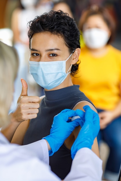 Retrato close-up tiro de paciente feliz do sexo feminino usa máscara facial sentar olhar para câmera mostrar polegar enquanto na fila de vacinação receber injeção de vacina tiro de agulha de seringa pelo médico.