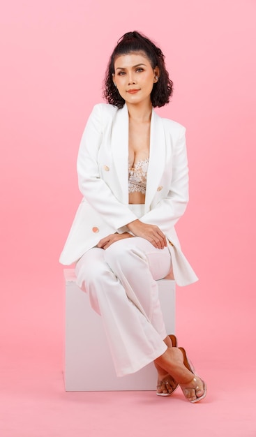 Retrato closeup estúdio tiro asiático jovem penteado encaracolado sexy empresária de sucesso profissional em moda branca terno casual e sutiã de renda sentado sorrindo pernas cruzadas posando no fundo rosa