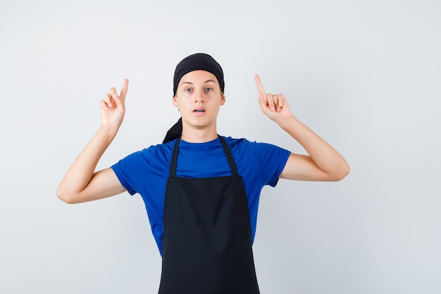 Retrato de cocinero adolescente masculino apuntando hacia arriba en camiseta, delantal y mirando sorprendido vista frontal