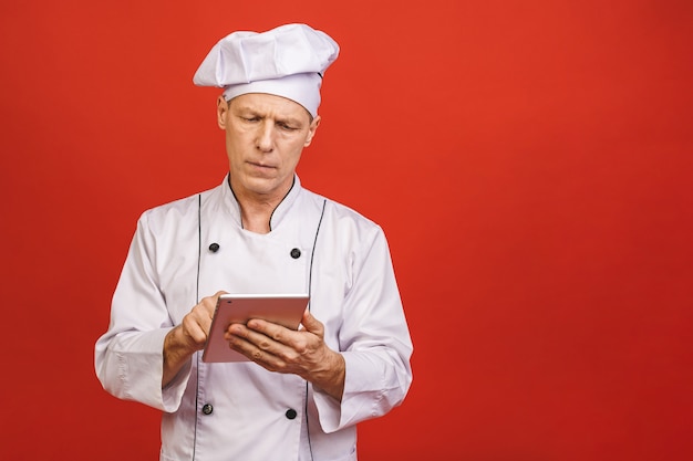 Retrato de un cocinero sonriente mayor serio del cocinero que sostiene la tableta aislada en un fondo rojo.