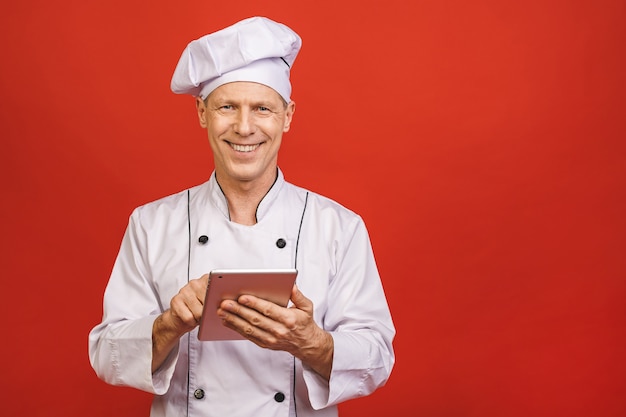 Retrato de un cocinero sonriente mayor sonriente del cocinero que sostiene la tableta aislada en un fondo rojo.