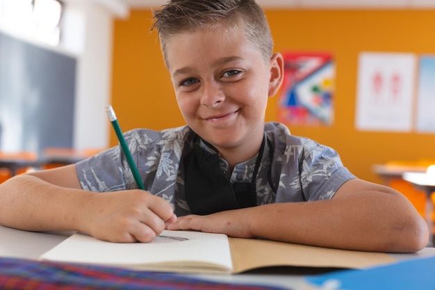 Foto retrato de un colegial caucásico sonriente con mascarilla sentado en el aula, escribiendo en el libro escolar