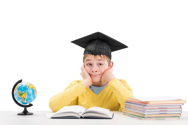 Retrato de colegial con sombrero de estudiante en la mesa junto a cuadernos y globo El niño hace su tarea sentado en su escritorio Aislado sobre fondo blanco
