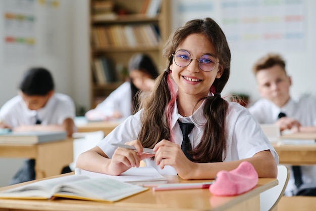 Retrato de una colegiala con anteojos sonriendo a la cámara mientras se sienta en su escritorio en la lección