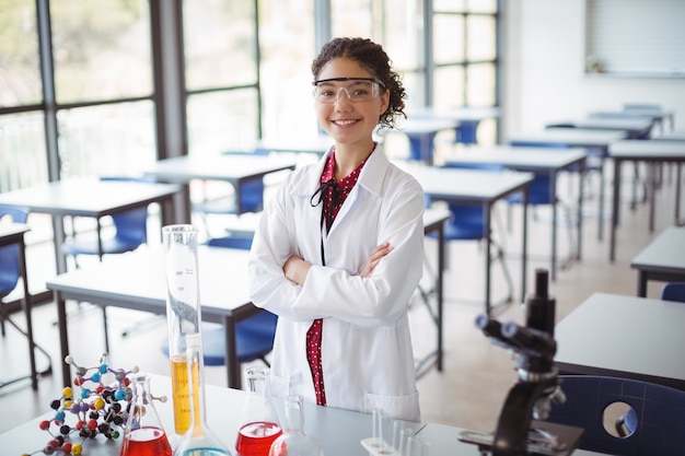 Retrato de colegiala en bata de laboratorio de pie con los brazos cruzados en el laboratorio